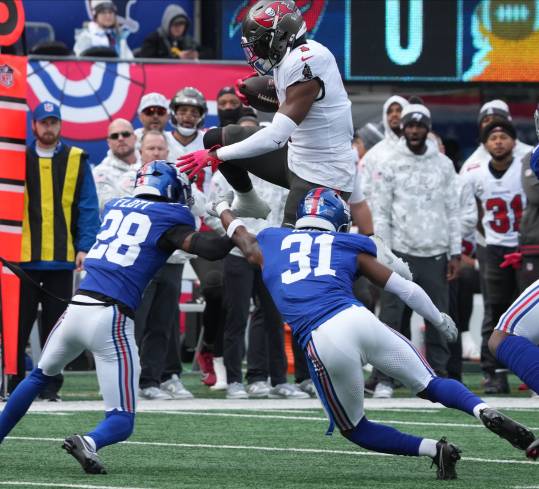 East Rutherford, NJ -- November 24, 2024 -- Rachaad White of Tampa Bay jumps over Cordale Flott and Tyler Nubin of the Giants in the first half as the Tampa Bay Buccaneers came to MetLife Stadium to play the New York Giants.